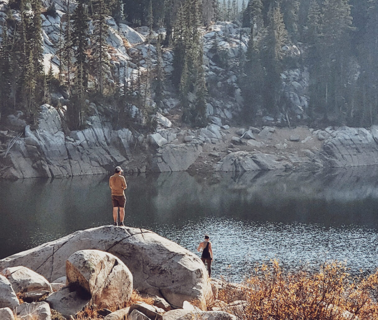 Bergsee mit Nadelbäumen. Im Vordergrund steht ein Mann auf einem Stein.