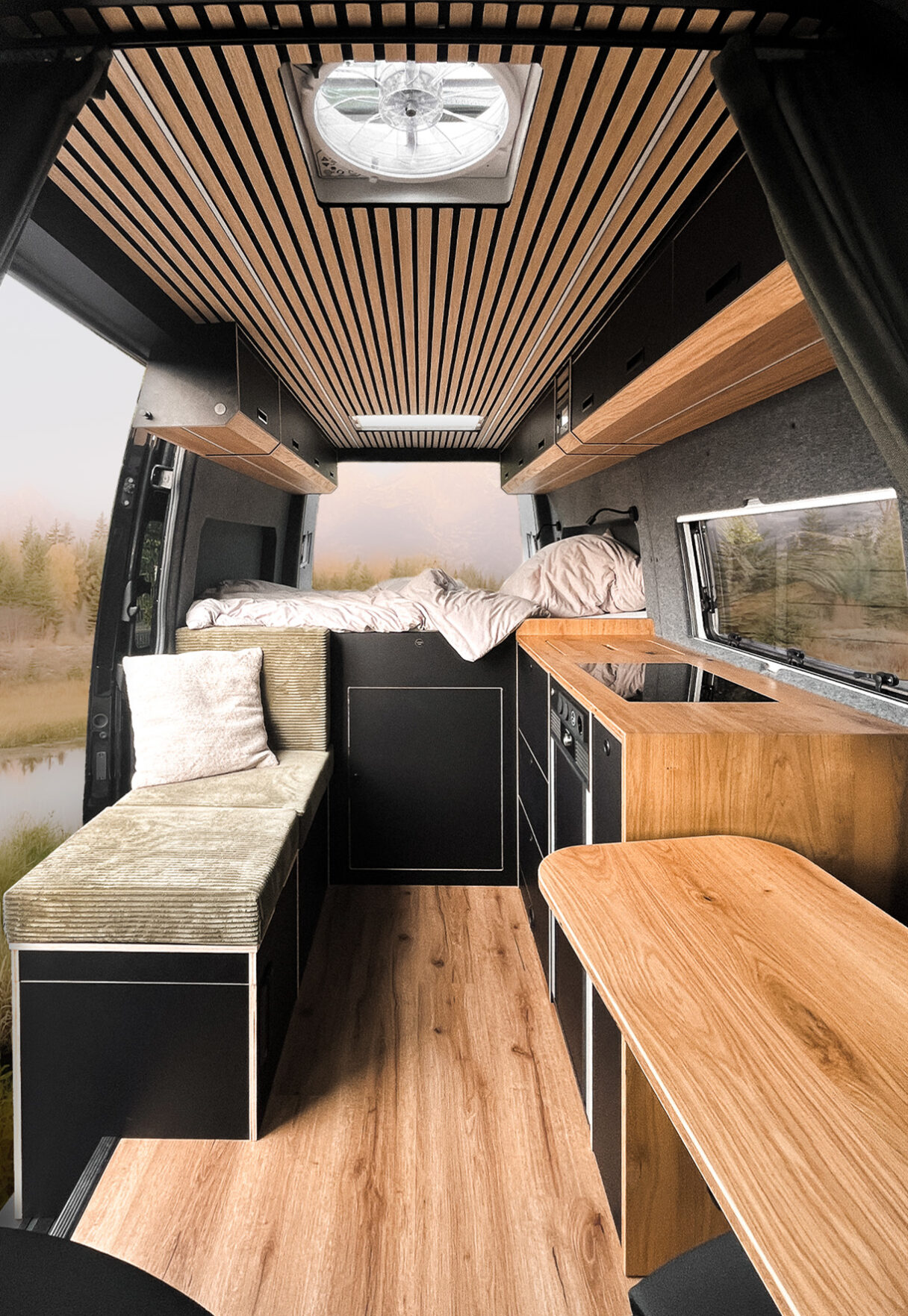 Innenraum eines zum Camper ausgebauten Van. Zu sehen ist ein Sitzbereich mit grünem Überzug, ein Kochbereich mit eingebautem Herd und ein Schlafbereich im Heck des Fahrzeuges. Die Optik ist in Holz und schwarz.