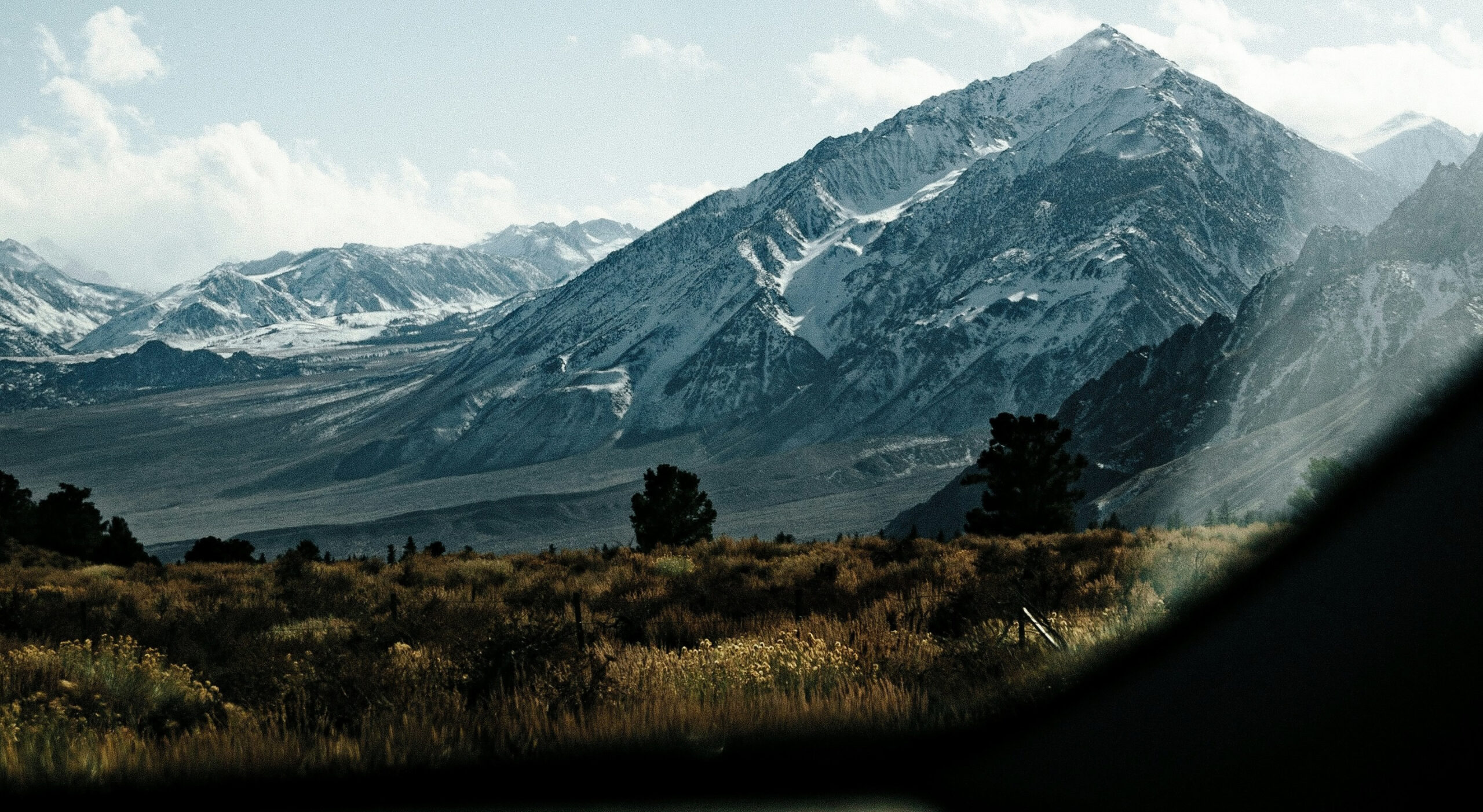 Blick aus der Frontscheibe eines Campers auf eine Berglandschaft. Im Vordergrund sind Wiesen, dahinter Berge, die mit Schnee bedeckt sind. Der Himmel ist hellblau und teilweise mit Wolken bedeckt.