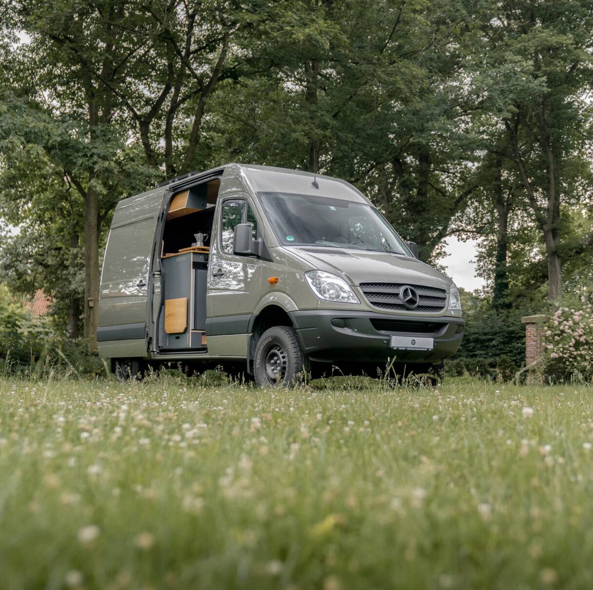 Mercedes Benz Sprinter in dunkelgrün steht auf einer Wiese vor einem Wald in der Natur. Die Schriebetür ist geöffnet und durch die Türe ist eine Küchenzeile zu sehen, die in den Van eingebaut wurde